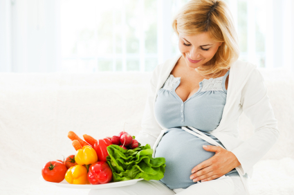 Cheerful beautiful pregnant woman sitting on the sofa with a bowl full of vegetables.
